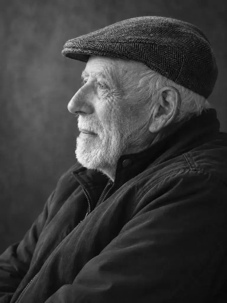 Portrait de profil en noir et blanc de Roger, coiffé d’une casquette et portant une barbe blanche, sur fond sombre